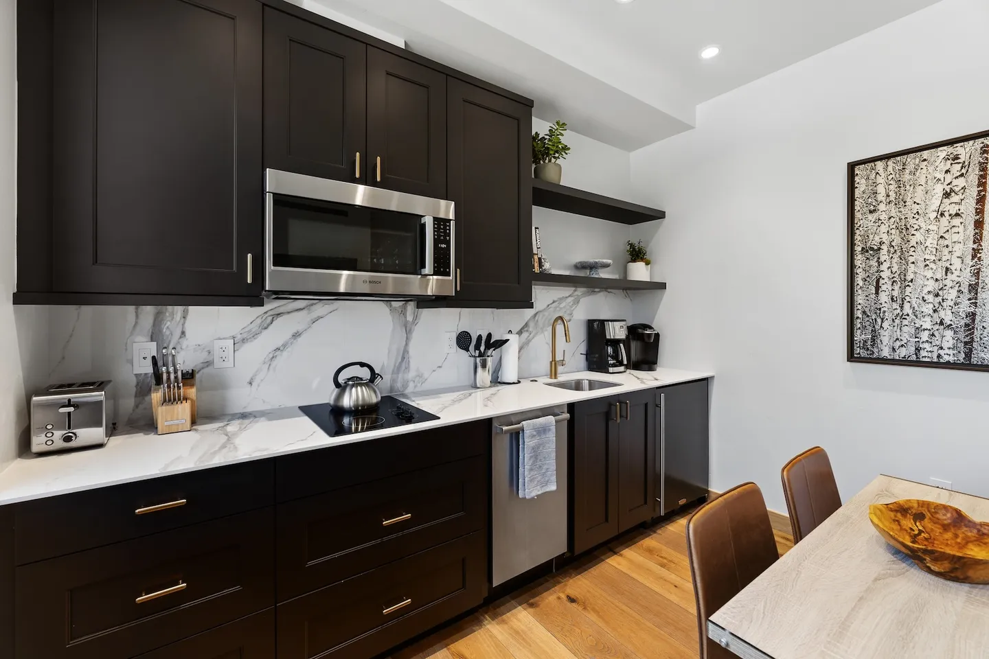 Modern kitchenette featuring dark cabinets, marble-patterned backsplash, stainless steel microwave, and small sink.