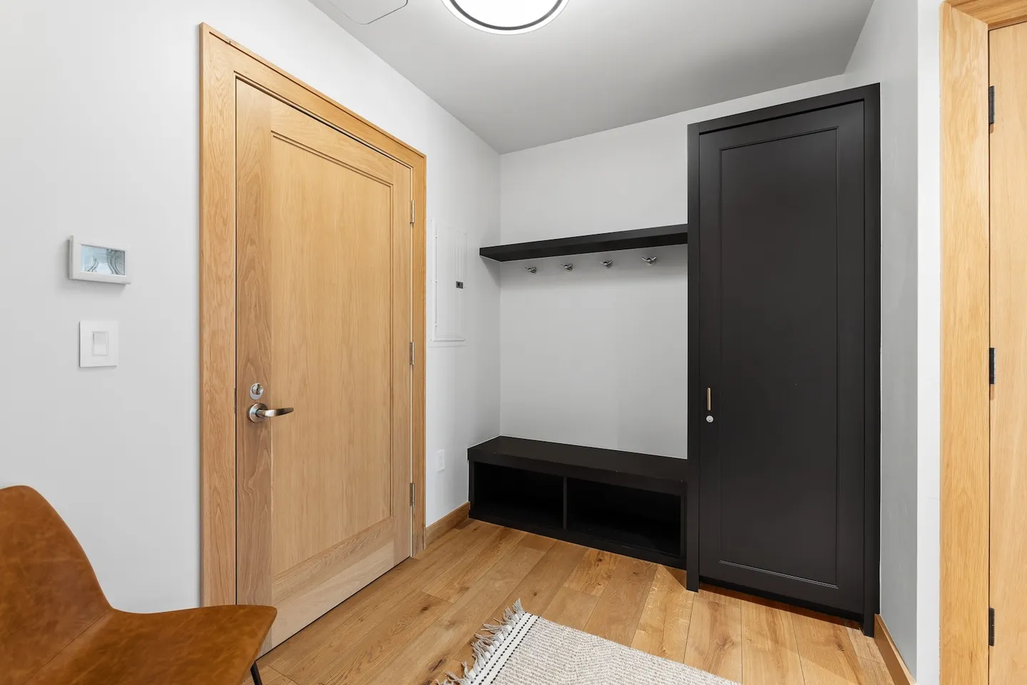 An entryway featuring light wood floors, a wooden interior door, and a built-in dark bench with coat hooks and a tall cabinet.