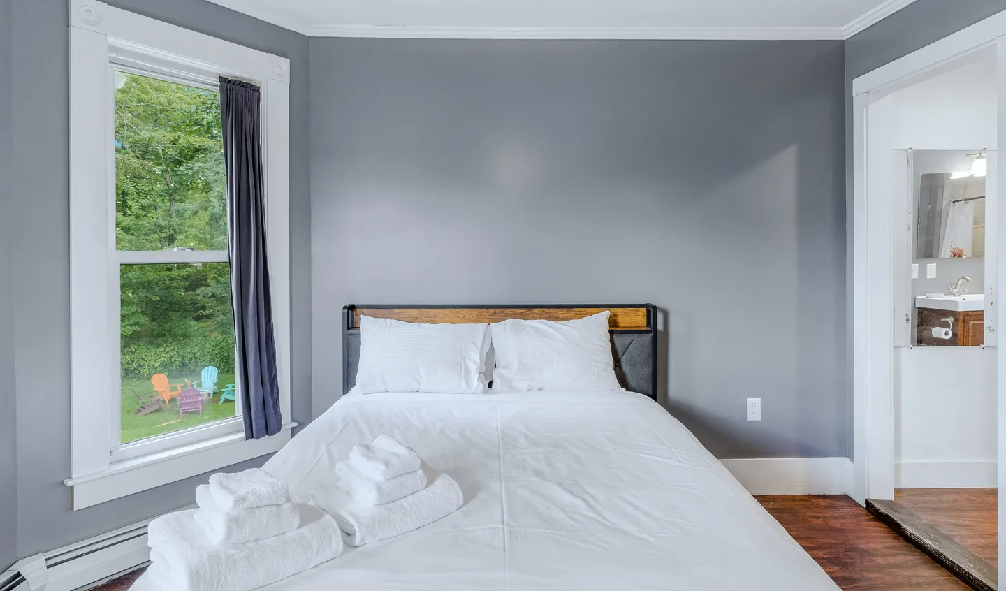 Bedroom with gray walls, a bed made with white linens and folded towels, a window overlooking the yard, and an open doorway to a bathroom.