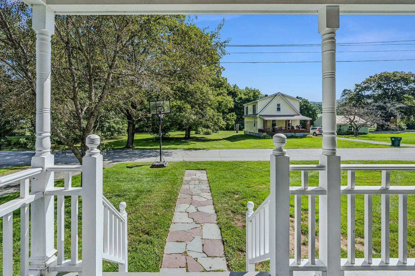 View looking out from a covered front porch with white railings and turned columns onto a stone walkway, grassy yard, and street.
