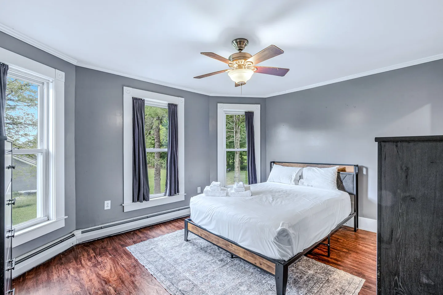 Bedroom with gray walls, wood-look flooring, multiple windows, a ceiling fan, and a made bed with white linens.