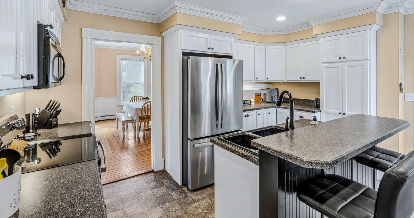 Kitchen with white cabinetry, dark countertops, a breakfast bar with black stools, and a stainless steel refrigerator next to a dining room doorway.