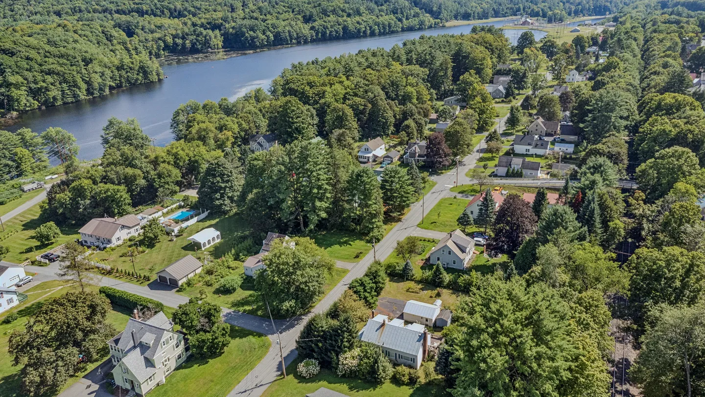 Aerial view of a neighborhood with houses, streets, dense green trees, and an adjacent river.