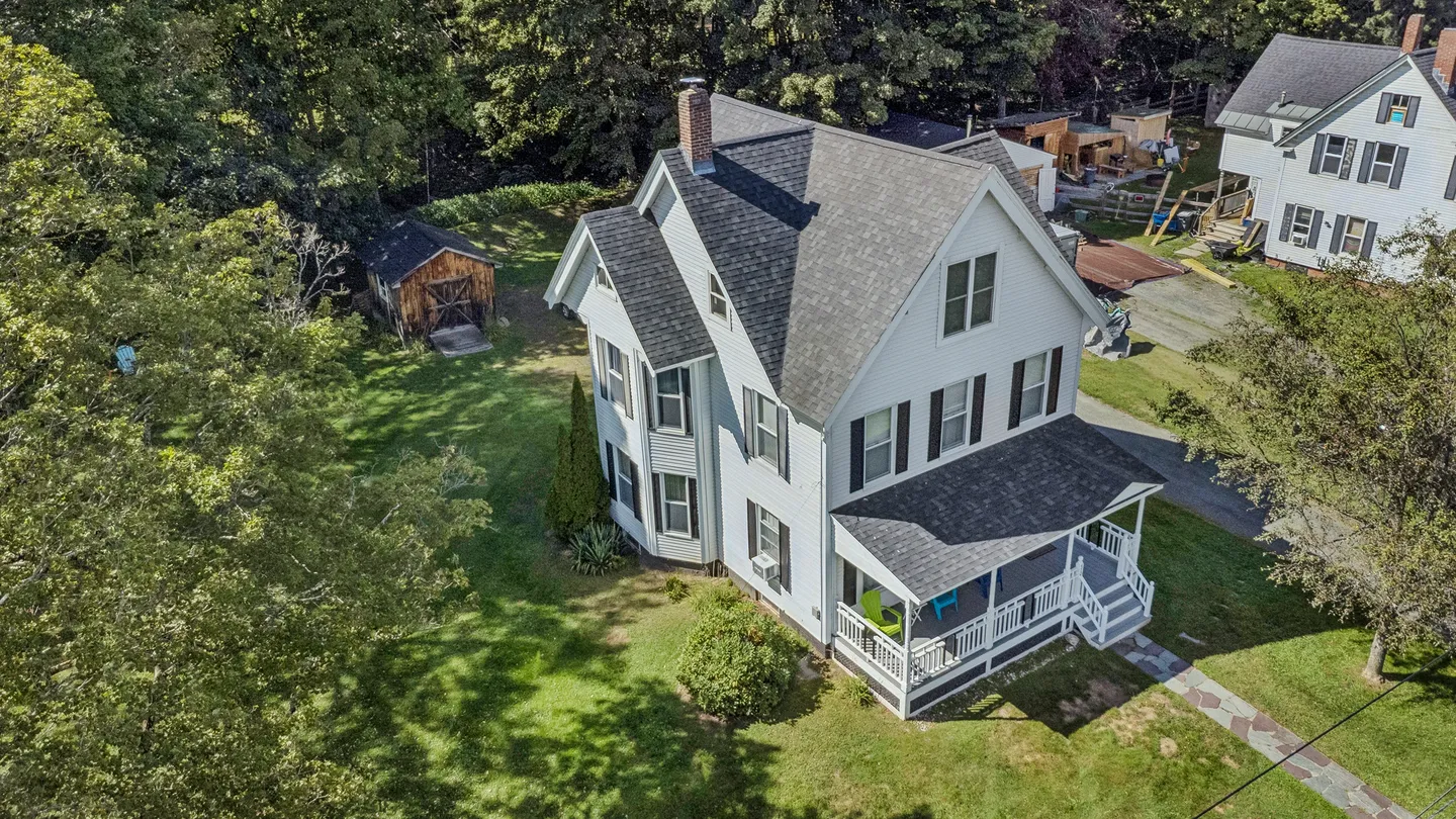 Aerial view of a multi-story house with light siding, dark shutters, a covered porch, and a grassy yard with trees.