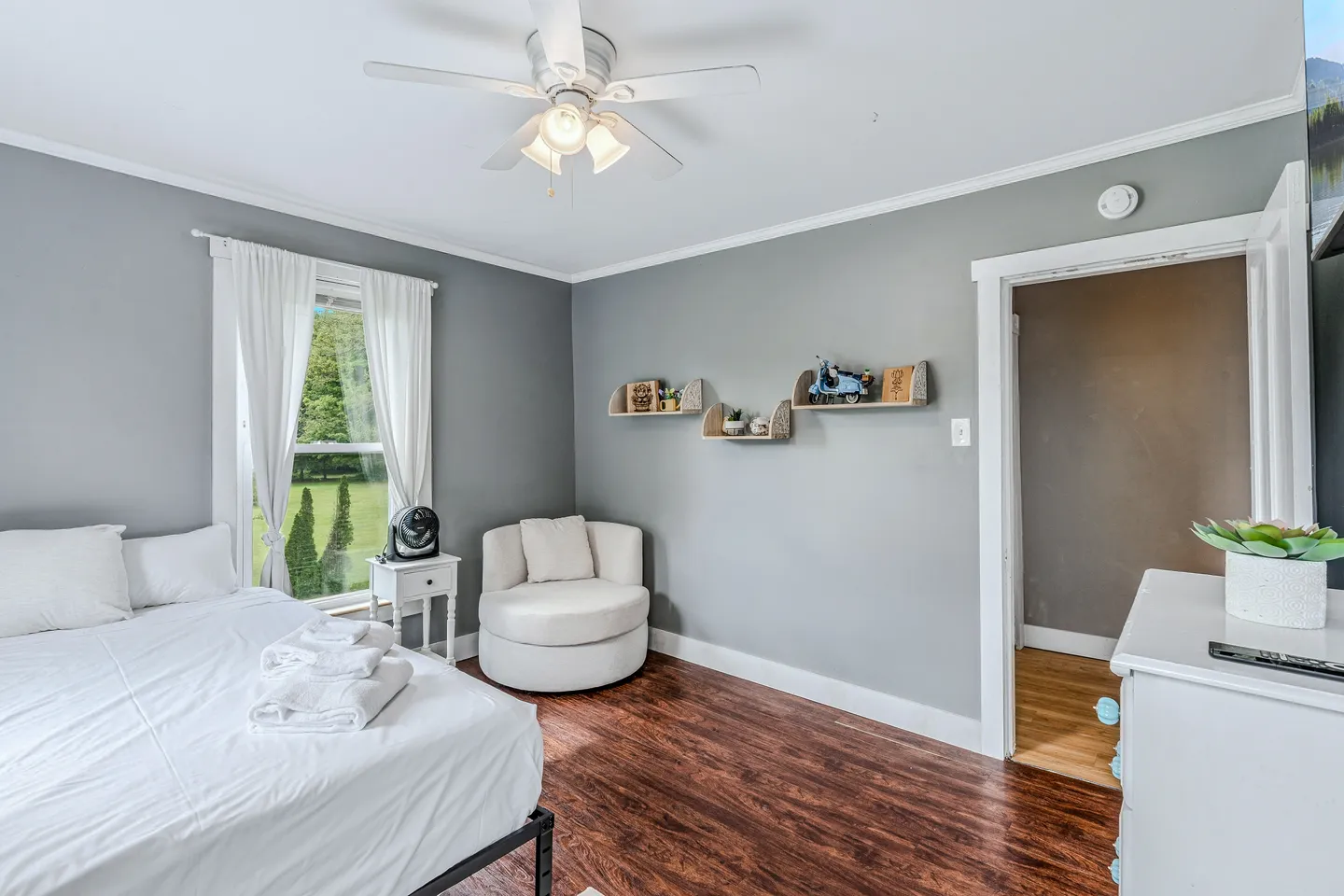 Bedroom with grey walls, dark wood-look flooring, a made bed with white linens, a swivel armchair, and wall shelves.