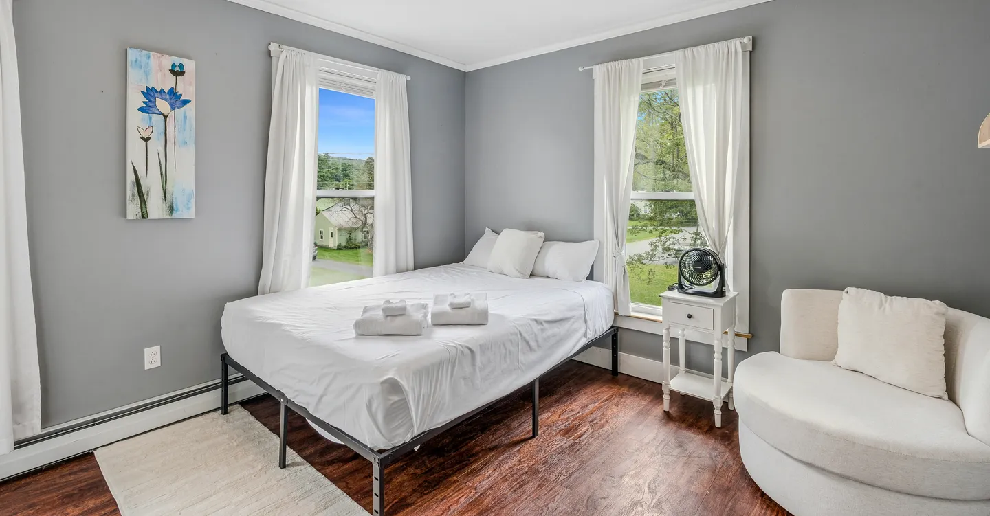 Bedroom with a metal-frame bed made with white linens, two windows with white curtains, a small bedside table, and a curved armchair.