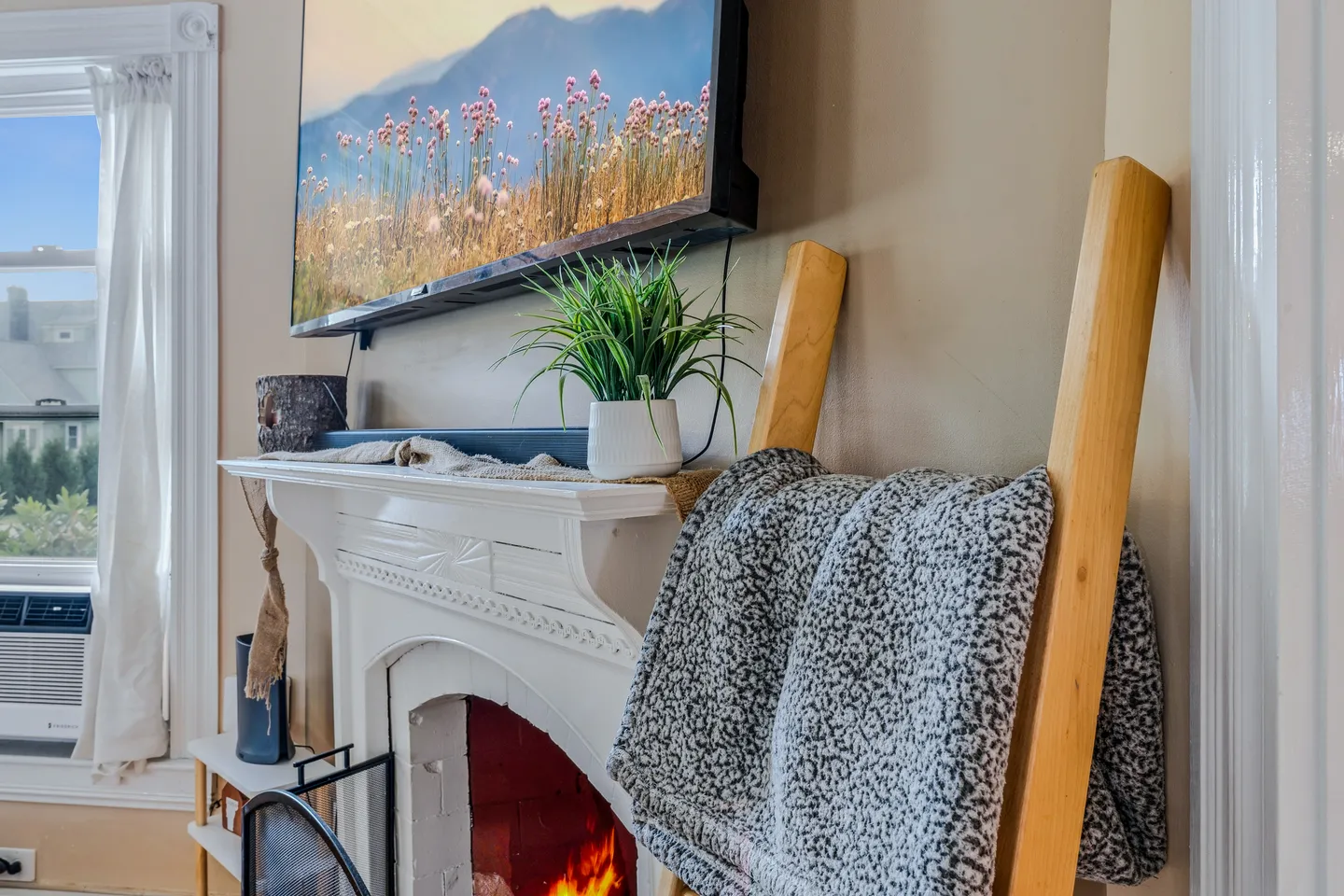 Close-up of a white painted fireplace mantel with a TV mounted above, a small potted plant, and a blanket ladder nearby.