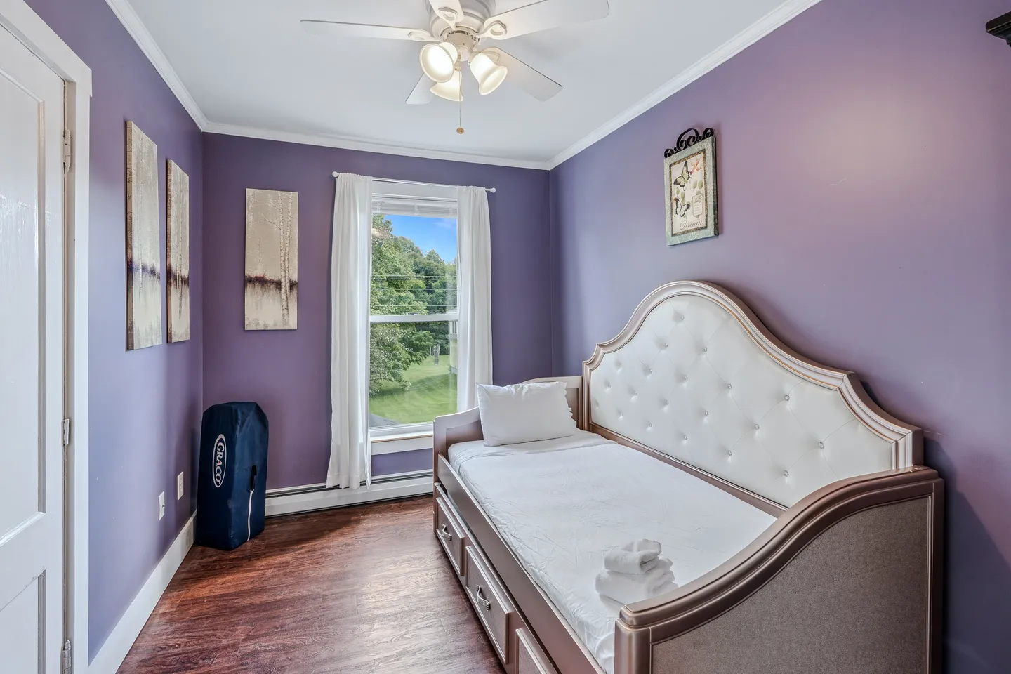 Bedroom with purple walls, dark wood-look flooring, a daybed with an upholstered tufted headboard, and a window overlooking greenery.