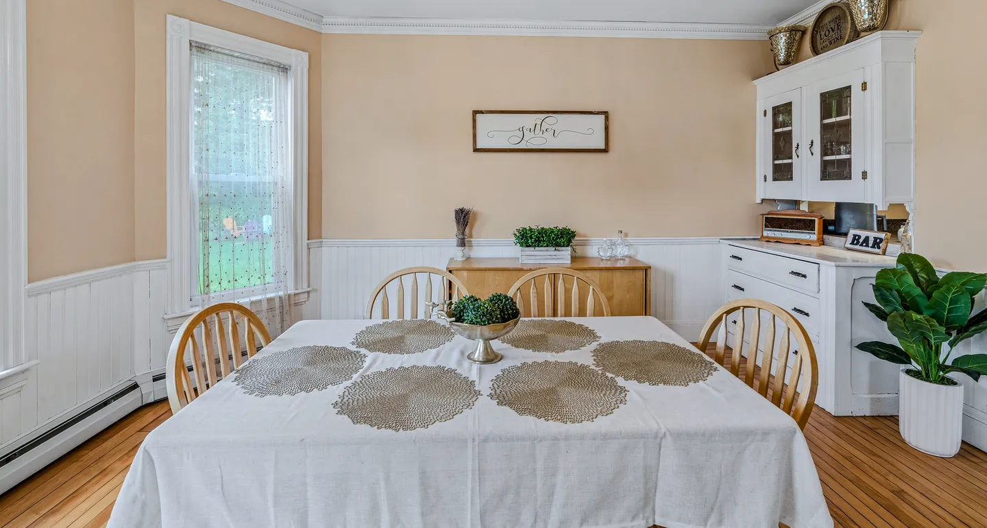 Dining room with a table set with placemats, wooden chairs, light peach walls with white wainscoting, and a white hutch cabinet.