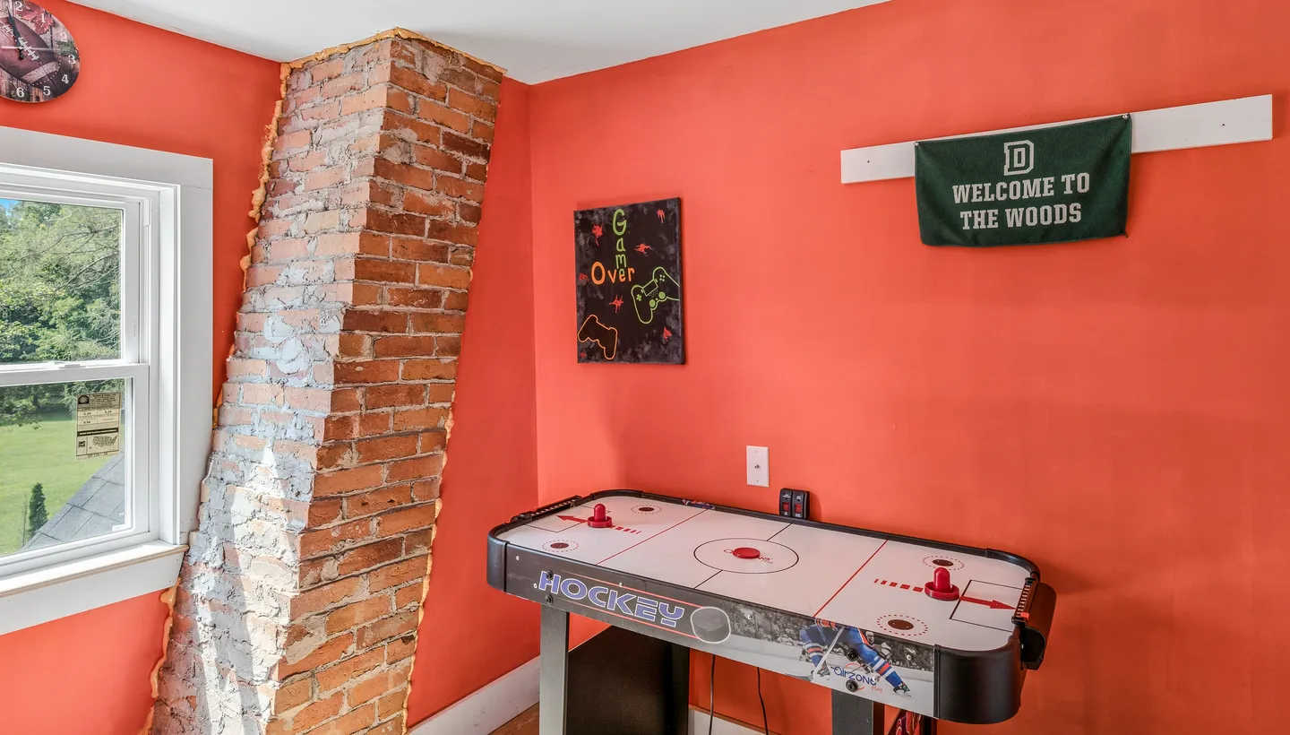 An air hockey table set against bright orange walls next to an exposed brick chimney and a window.