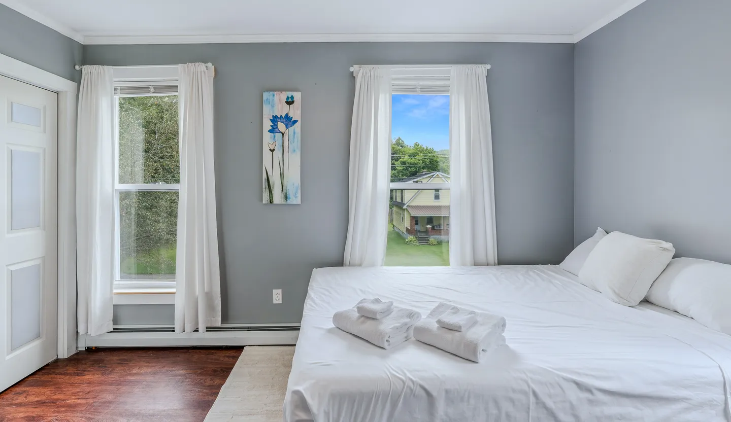 Bedroom with blue-gray walls, a bed made with white linens and folded towels, and two windows framed by white curtains.