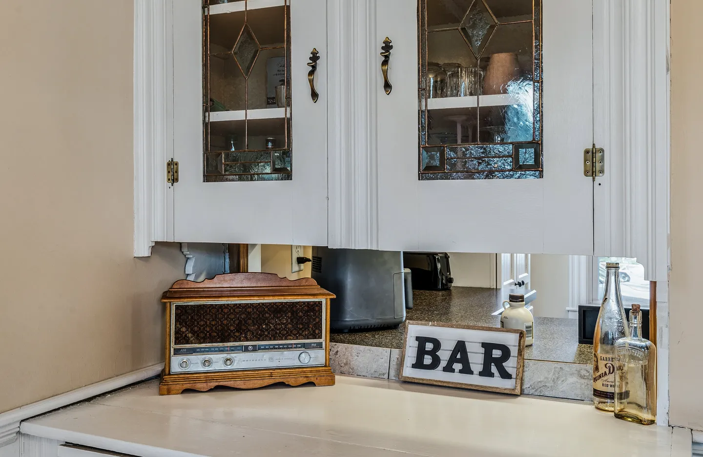 A pass-through bar counter beneath white glass-front cabinets featuring a vintage-style radio, bottles, and a BAR sign.