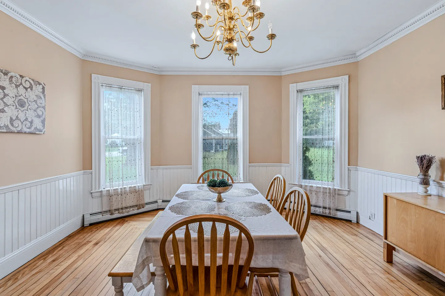 Dining room with wood flooring, three bay windows with lace curtains, and a chandelier hanging over a set dining table.