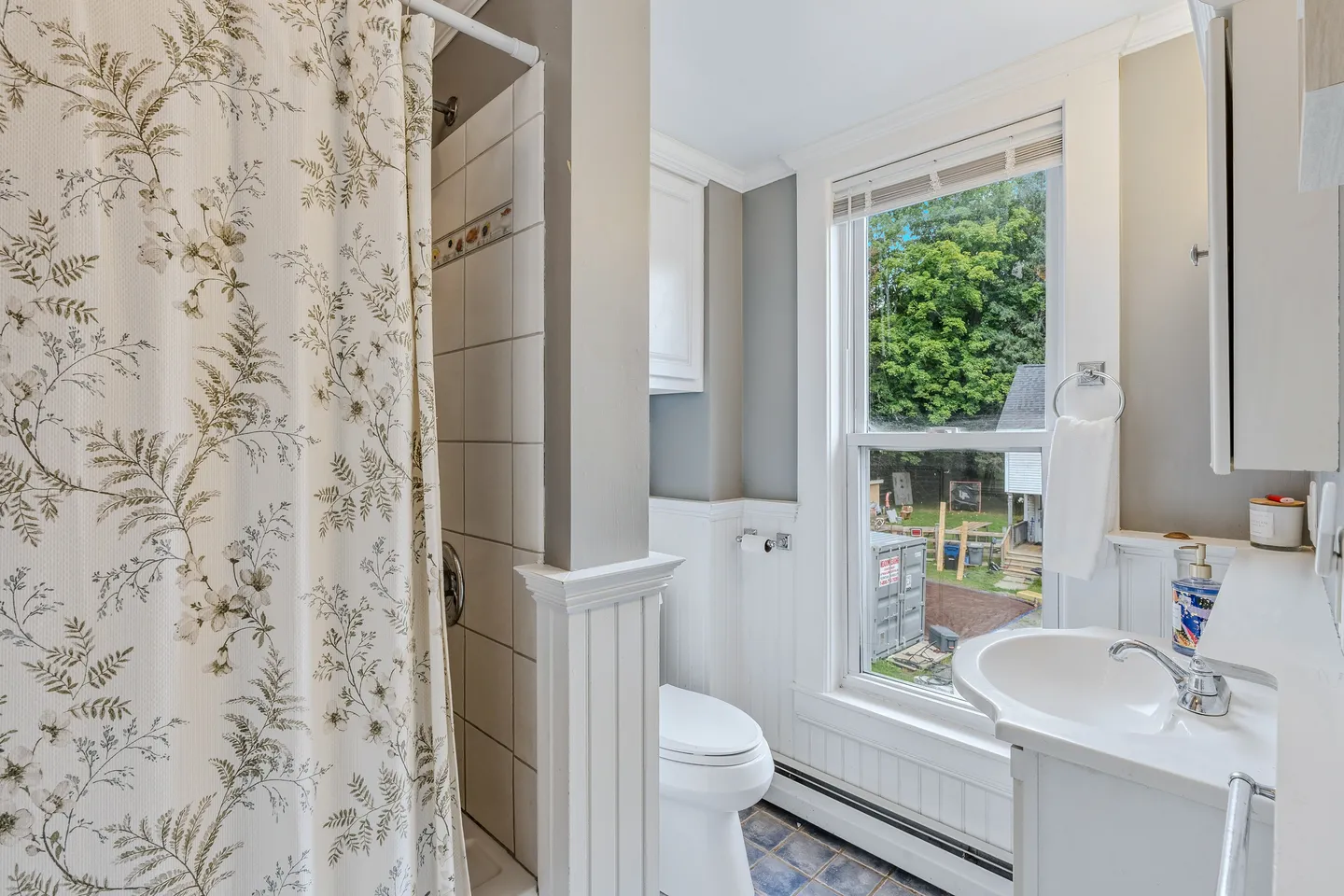 Bathroom with white wainscoting, shower with a floral curtain, white toilet, vanity sink, and window overlooking trees.