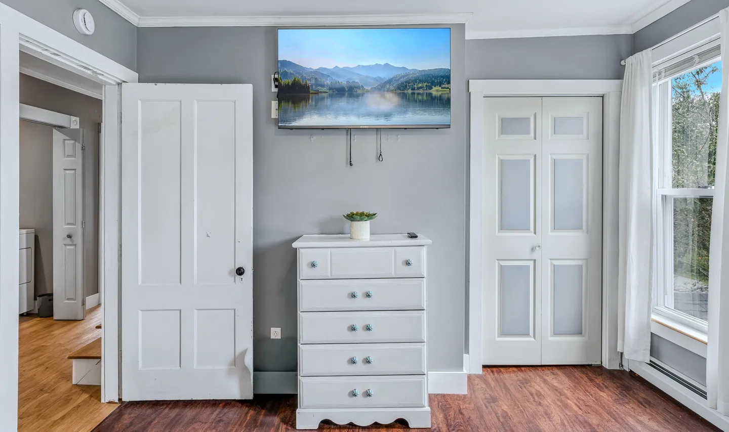 A white dresser sitting beneath a wall-mounted flat-screen television displaying a landscape photo, flanked by two white interior doors.