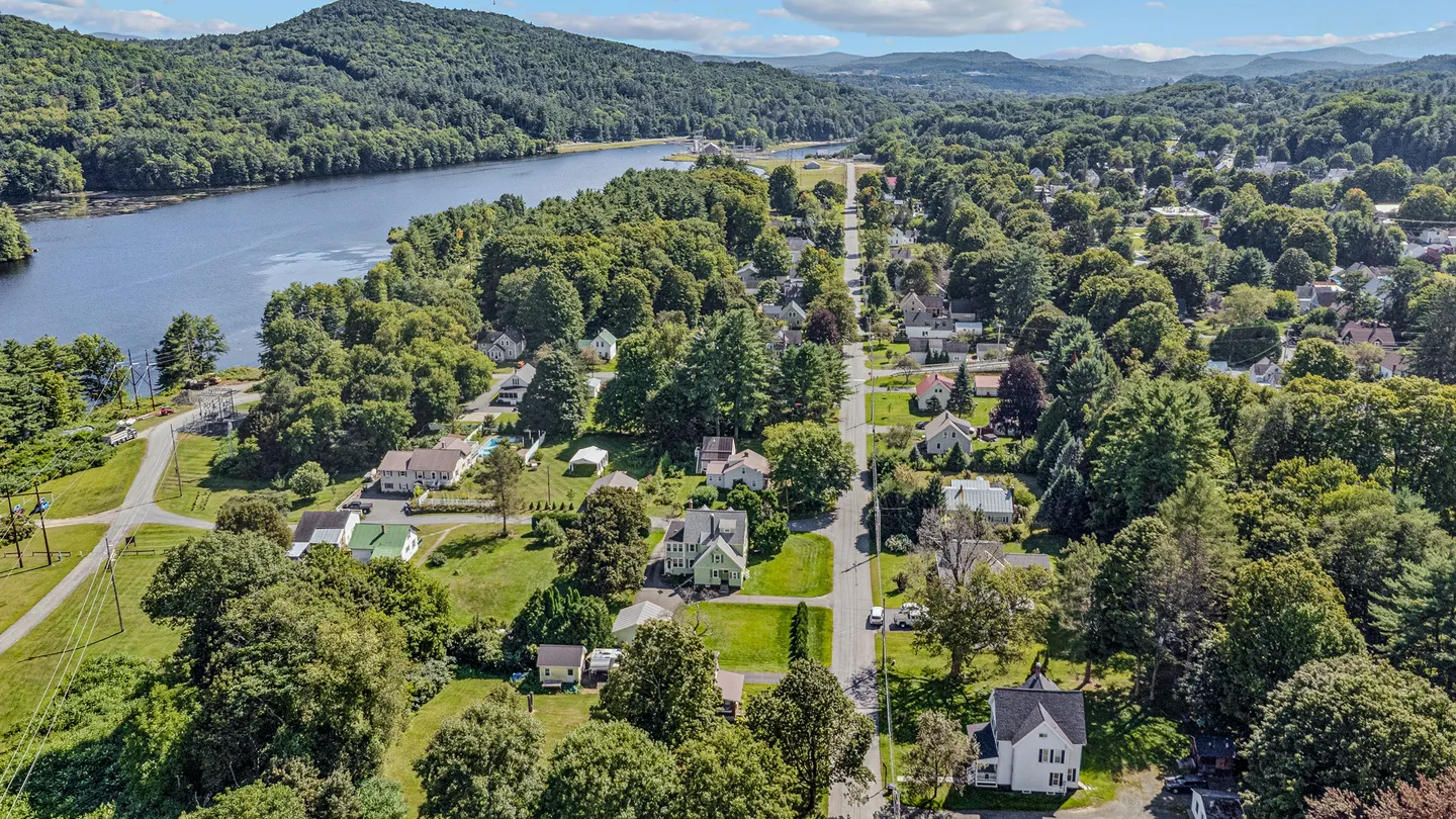 Aerial view of a neighborhood street lined with houses, dense green trees, and an adjacent wide river.