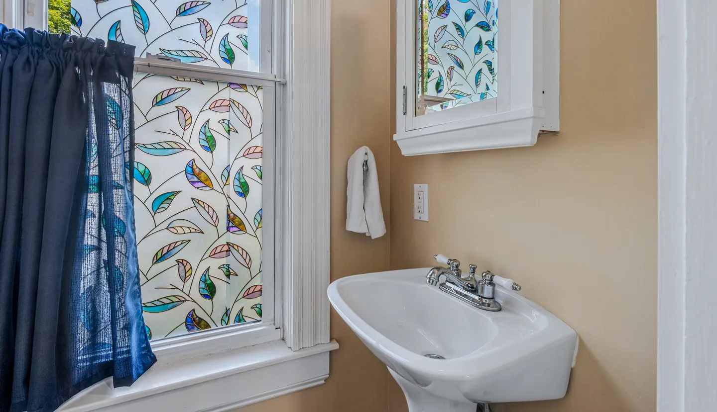 Pedestal sink in a bathroom beside a window with decorative patterned film and a dark curtain.