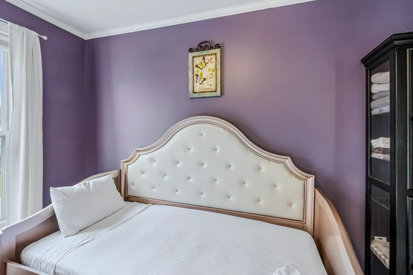 Bedroom corner with purple walls featuring a daybed with a tufted headboard and an adjacent tall black cabinet.