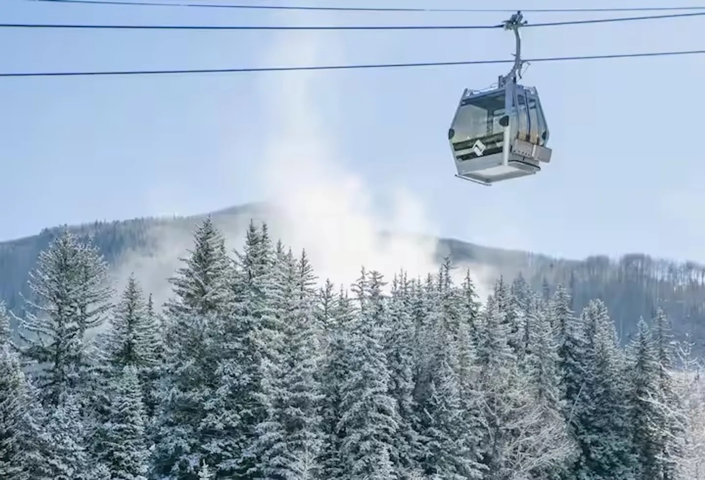 A ski gondola cabin suspended on overhead cables above snow-covered evergreen trees.