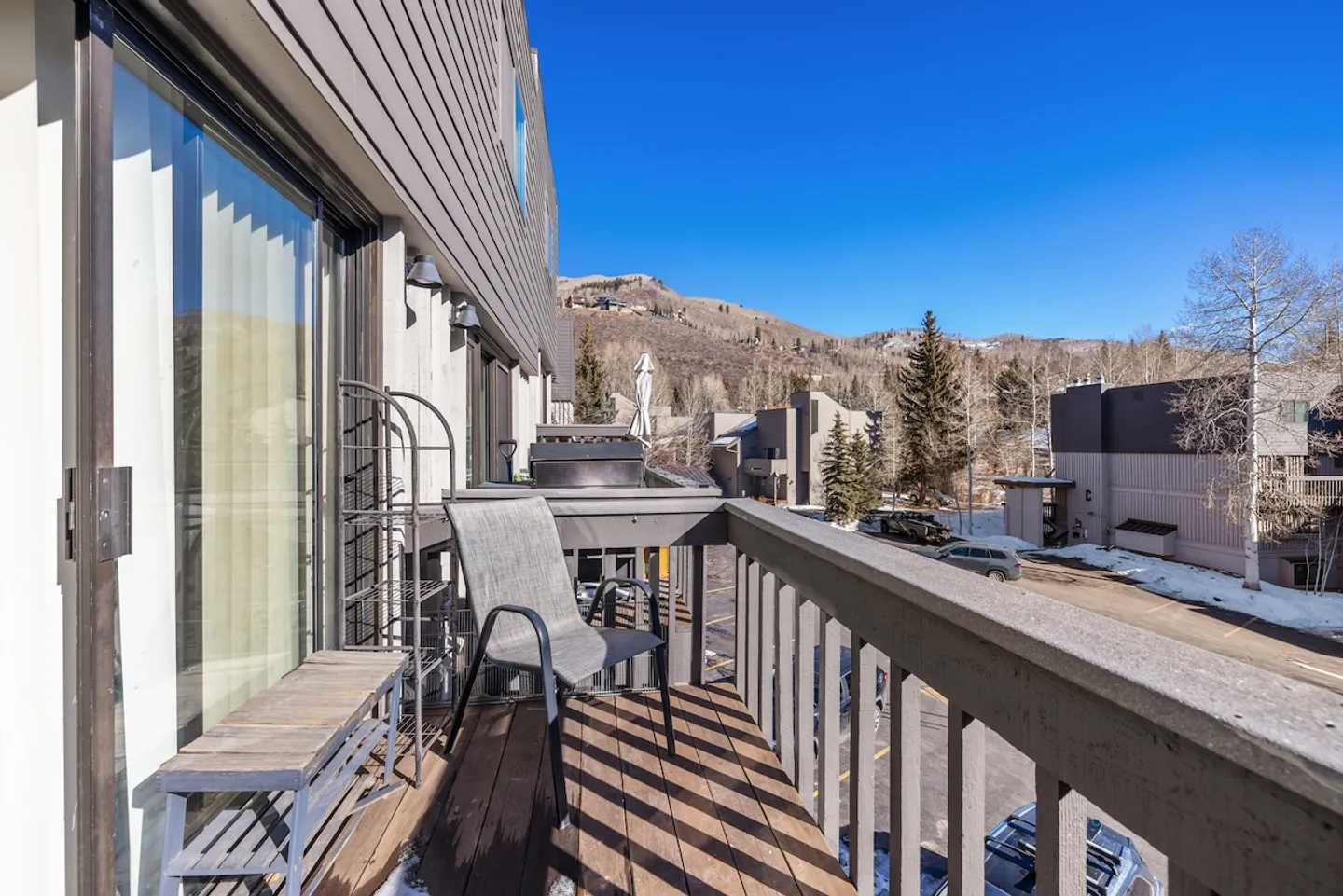 Balcony with wooden decking, railing, an armchair, and a small bench overlooking a parking area and distant mountains under a clear blue sky.