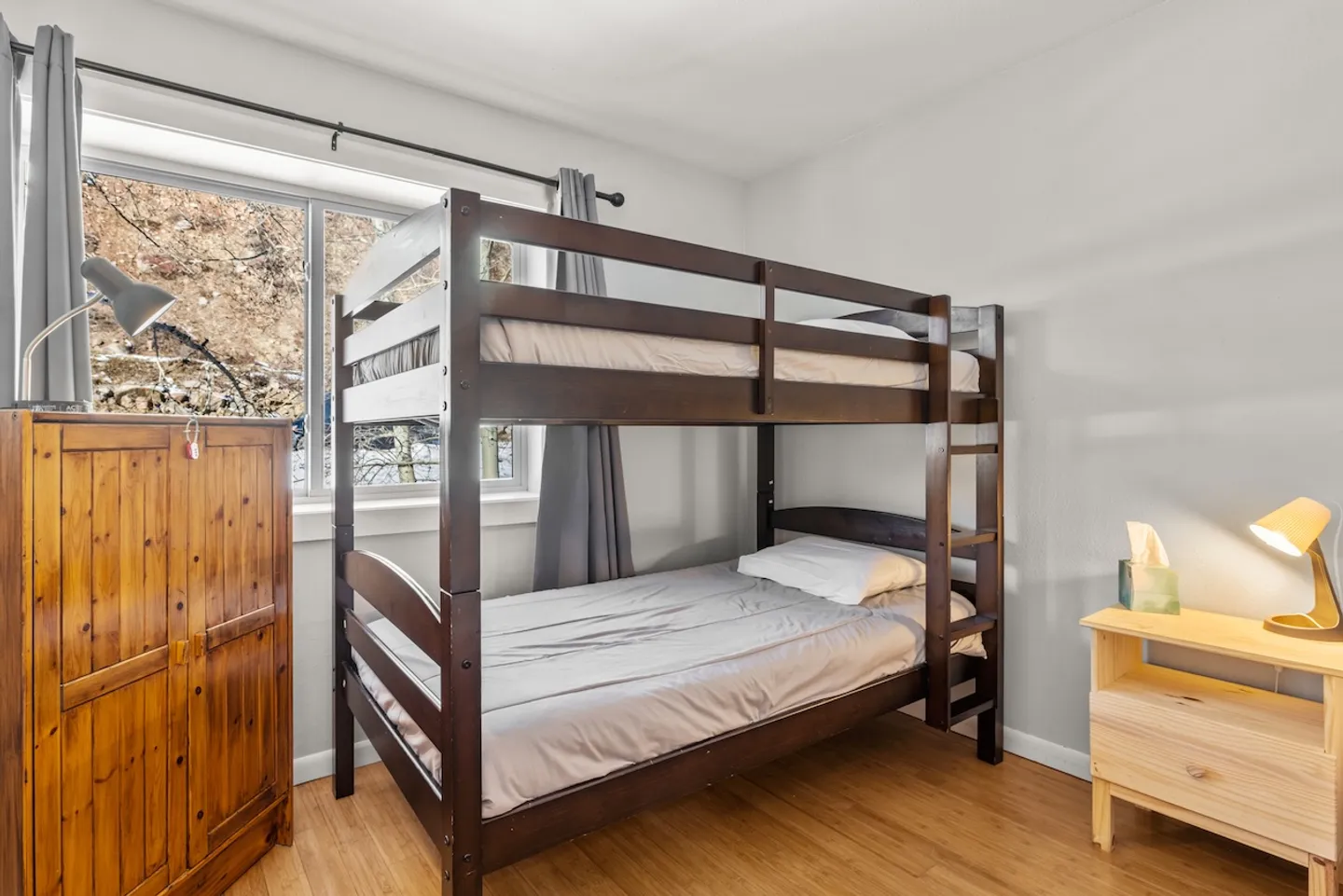 Bedroom featuring a dark wood bunk bed between a tall wooden wardrobe and a light wood nightstand with a lamp.