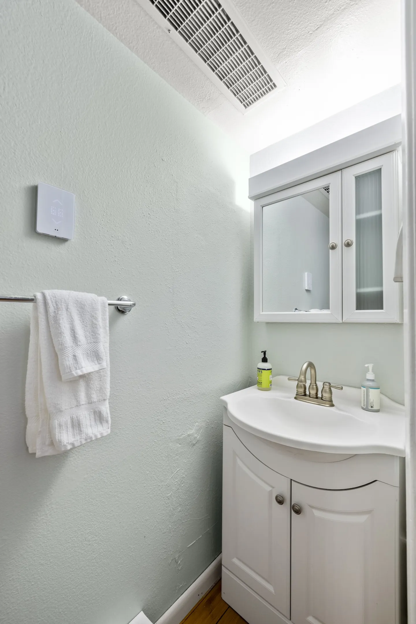 A compact bathroom vanity with a white curved cabinet, integrated sink, mirrored medicine cabinet, and a towel bar on the pale wall.