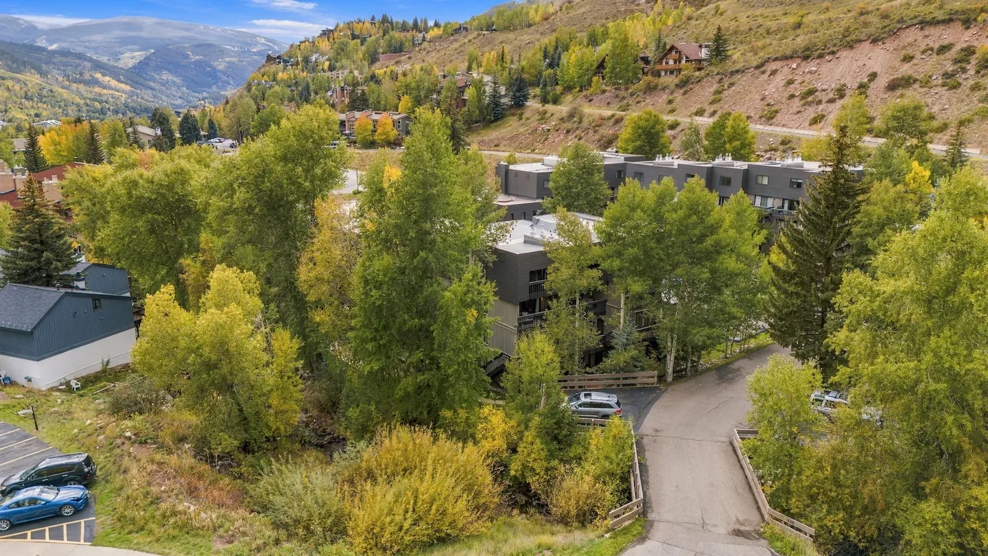 Aerial view of a multi-unit residential building surrounded by green and yellow trees against a mountain hillside.