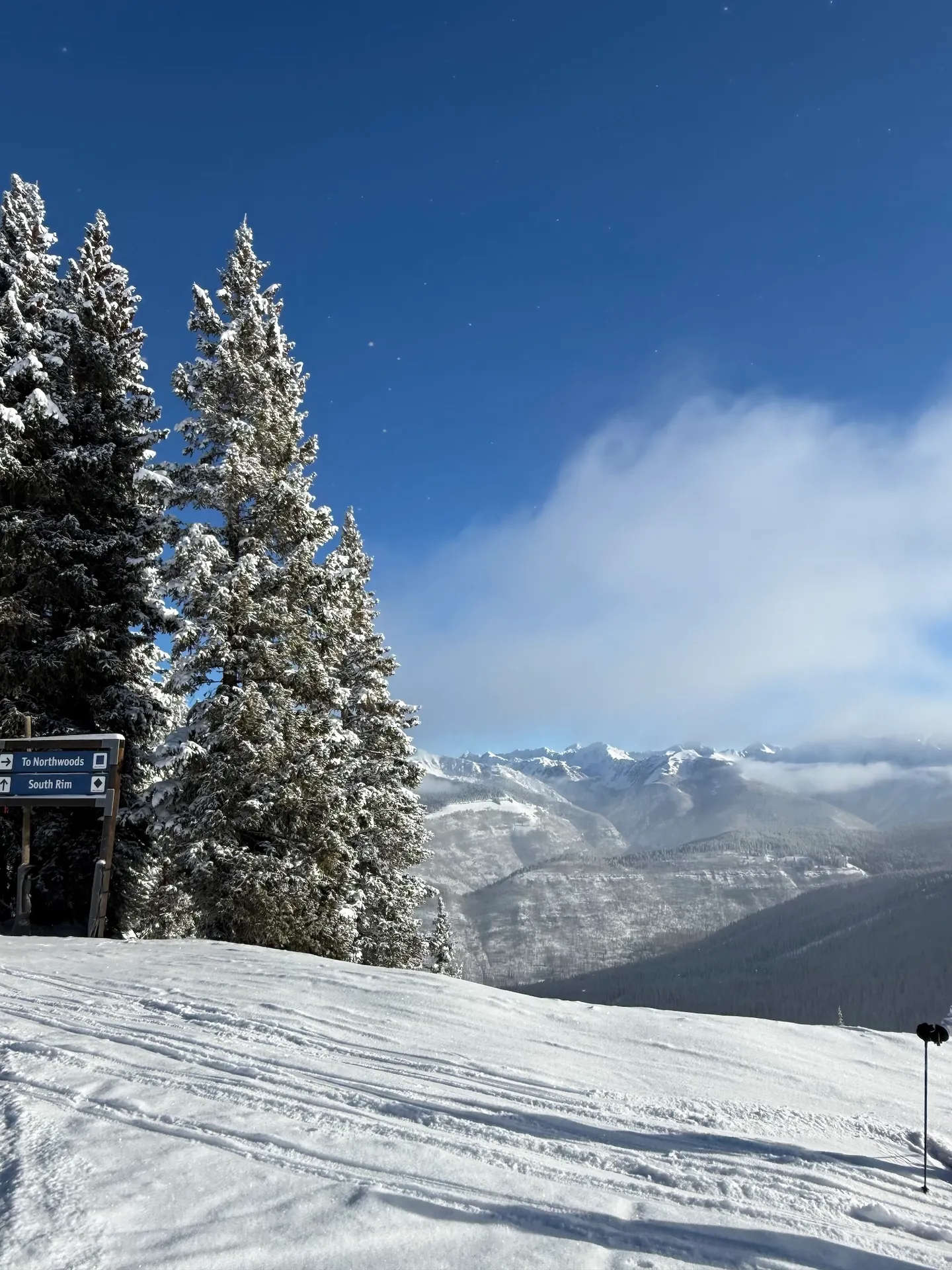 A snow-covered ski trail with evergreen trees, trail signs, and a wide panoramic view of distant mountains under a blue sky.