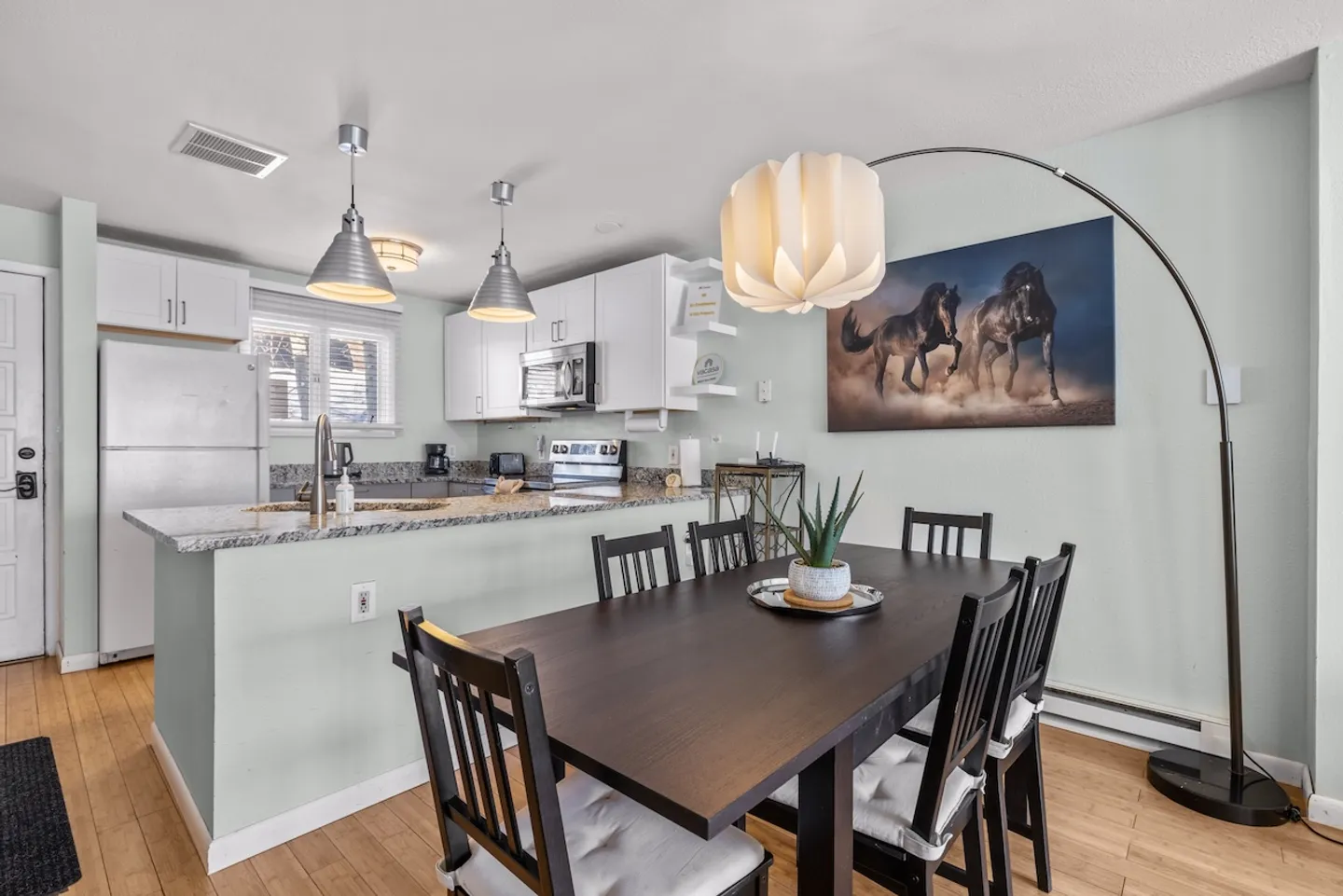 Dining area with a dark wood table and chairs situated adjacent to an open kitchen with white cabinets and granite-look counters.