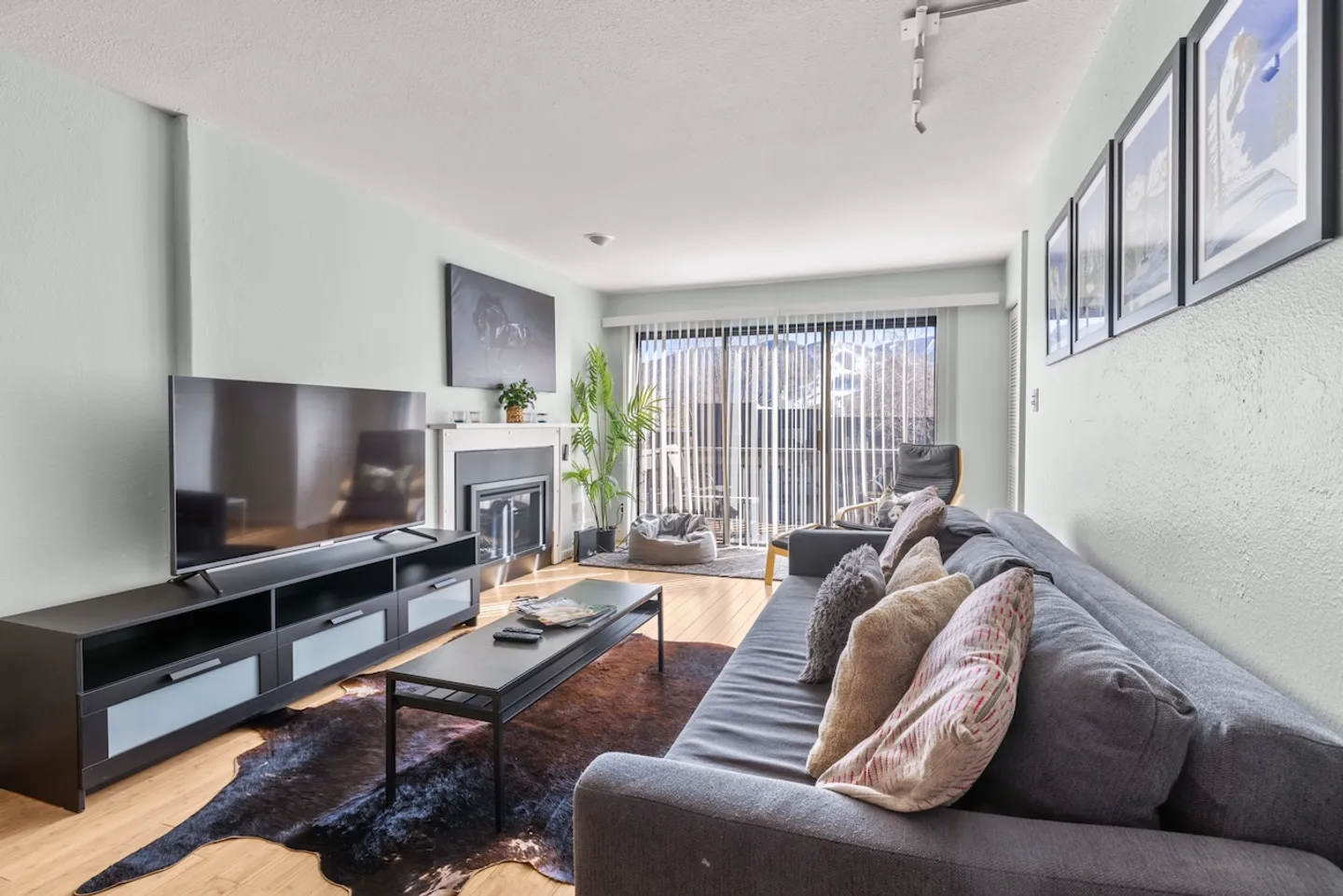 Living room with a gray sofa, dark media console with TV, fireplace, coffee table over an animal hide rug, and sliding glass doors leading outside.