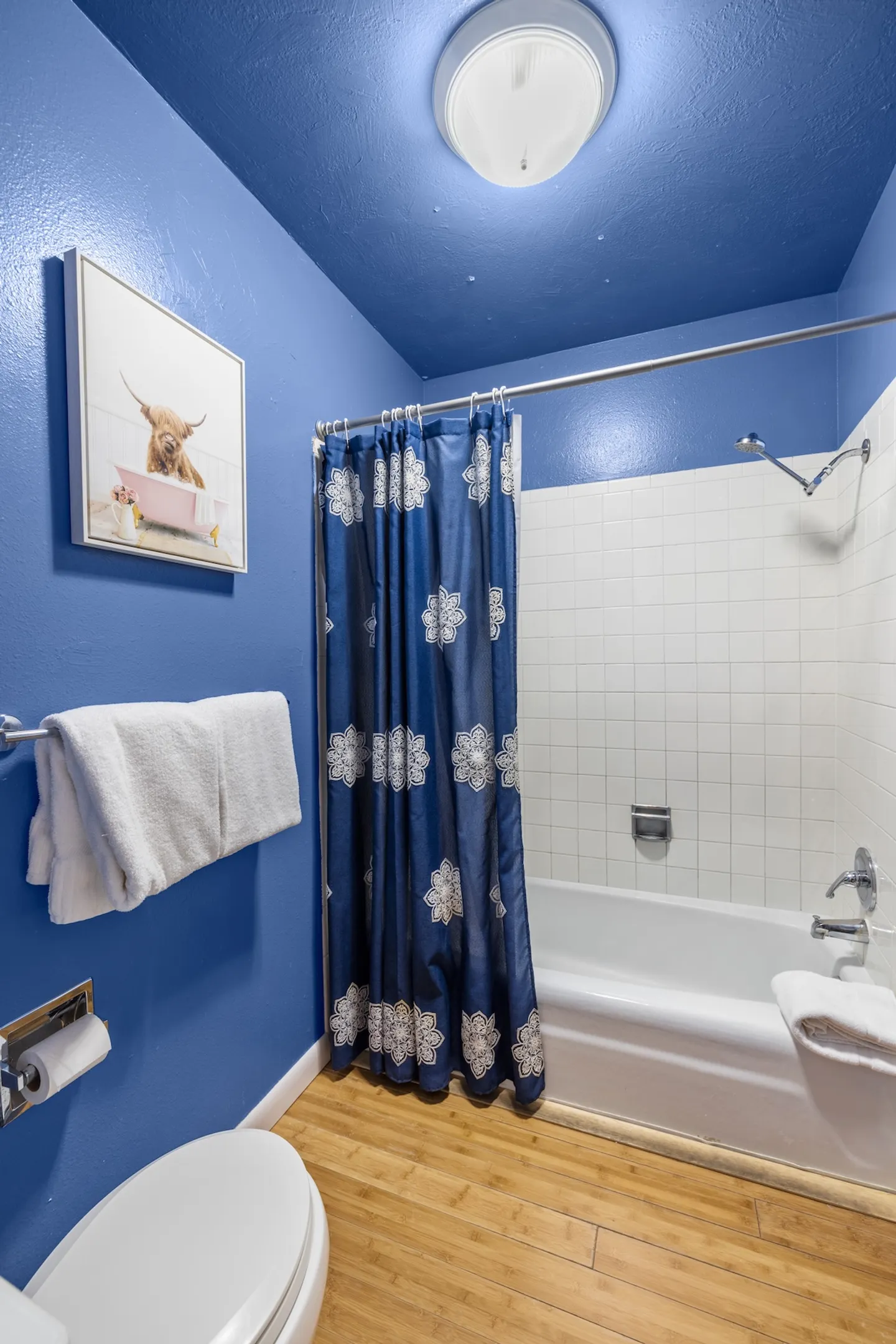 Bathroom with vibrant blue walls, white-tiled bathtub-shower combination, patterned blue shower curtain, and wood-look flooring.