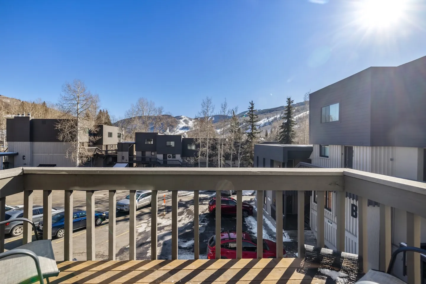 View from an outdoor balcony overlooking a parking lot, neighboring multi-unit buildings, and snow-covered mountains under a bright sunny sky.
