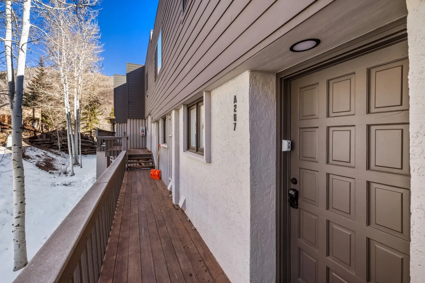 Exterior covered walkway and entry door of unit A207 alongside a snowy hillside with bare birch trees.