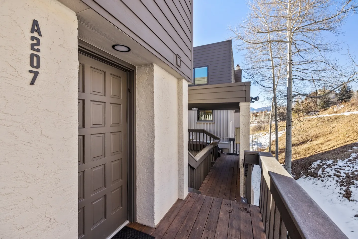 Exterior entrance to unit A207 showing a brown paneled front door along a wooden walkway and railing with snowy hillside in the background.