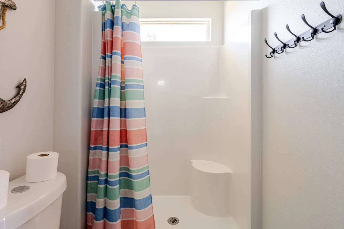 A bathroom stall shower with a colorful striped curtain, built-in corner seat, toilet to the left, and wall-mounted coat hooks to the right.