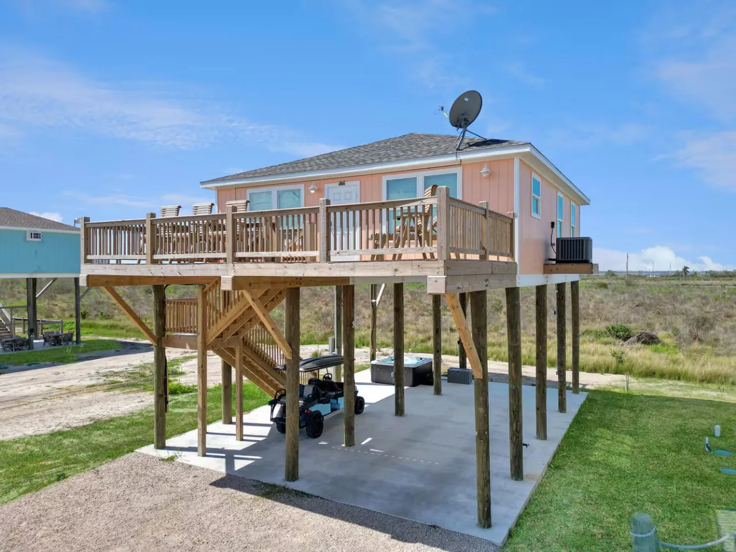 Exterior of an elevated peach stilt house with a wooden deck, covered ground-level parking, a golf cart, and a hot tub under a bright blue sky.