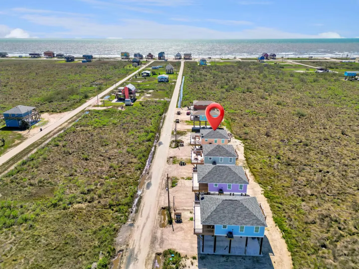 Aerial view of a coastal neighborhood with stilt houses leading toward the ocean, highlighted by a red location pin.