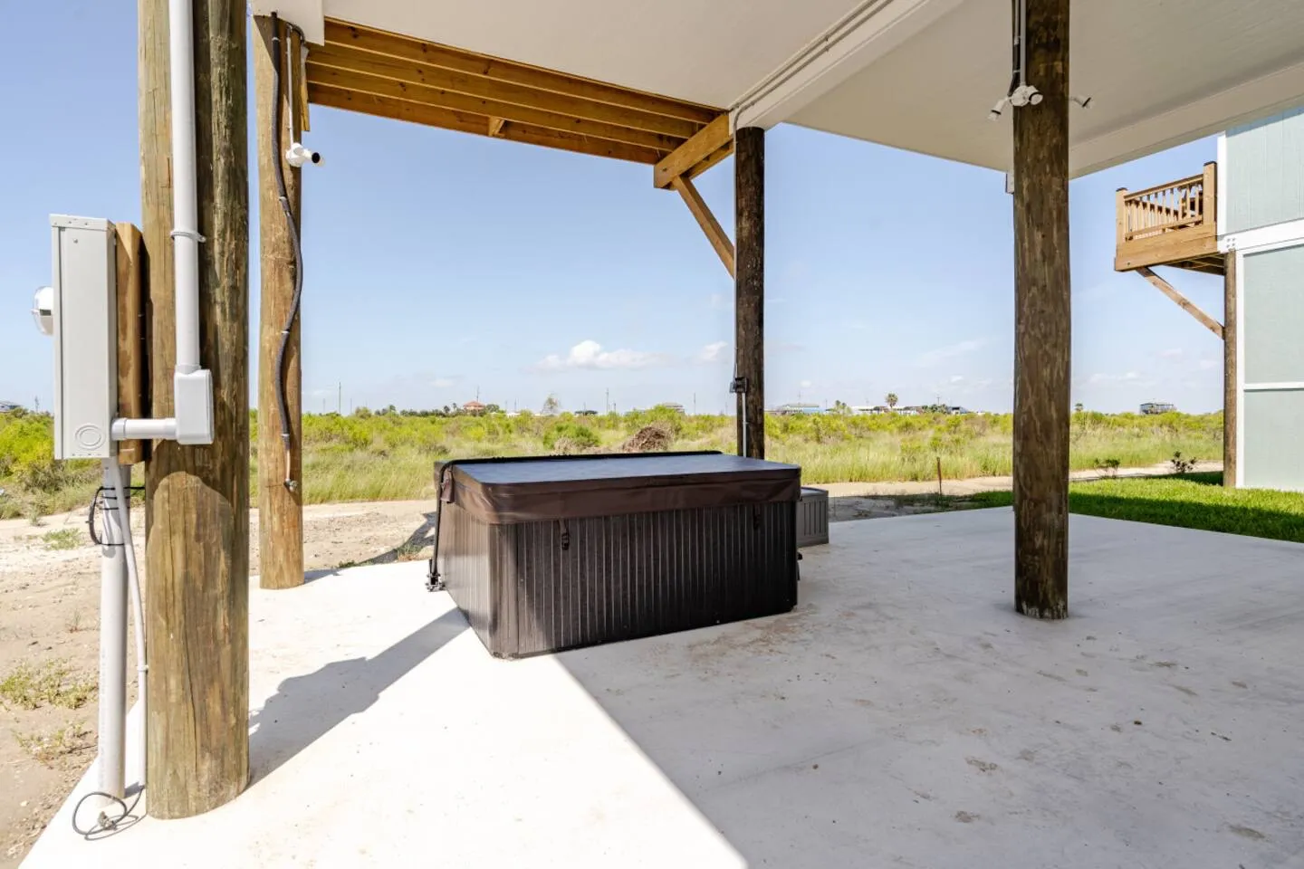 A covered hot tub on a concrete patio beneath a stilt home, looking out over coastal grassy fields.