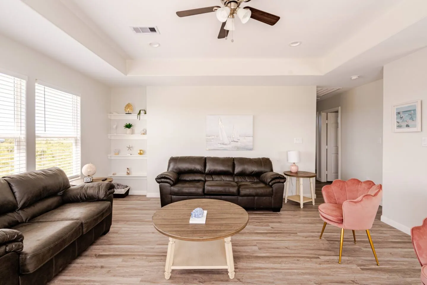 Living room with two dark leather sofas, a round wooden coffee table, a pink scalloped armchair, and light wood-plank flooring under a ceiling fan.