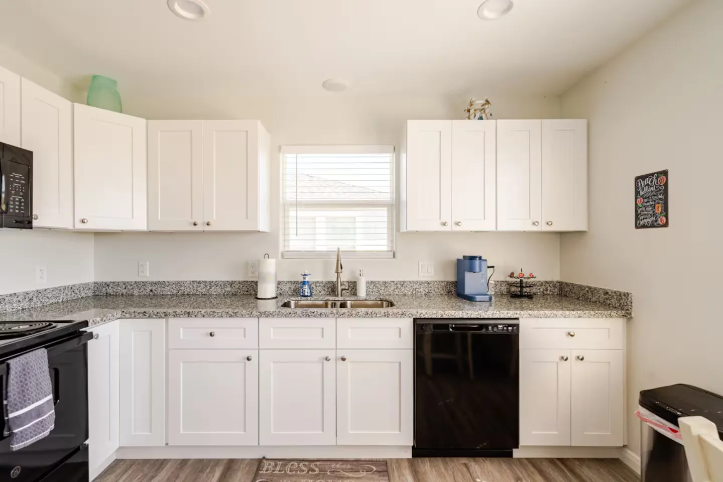 Kitchen with white shaker cabinets, speckled granite countertops, a double sink below a window, and black appliances.