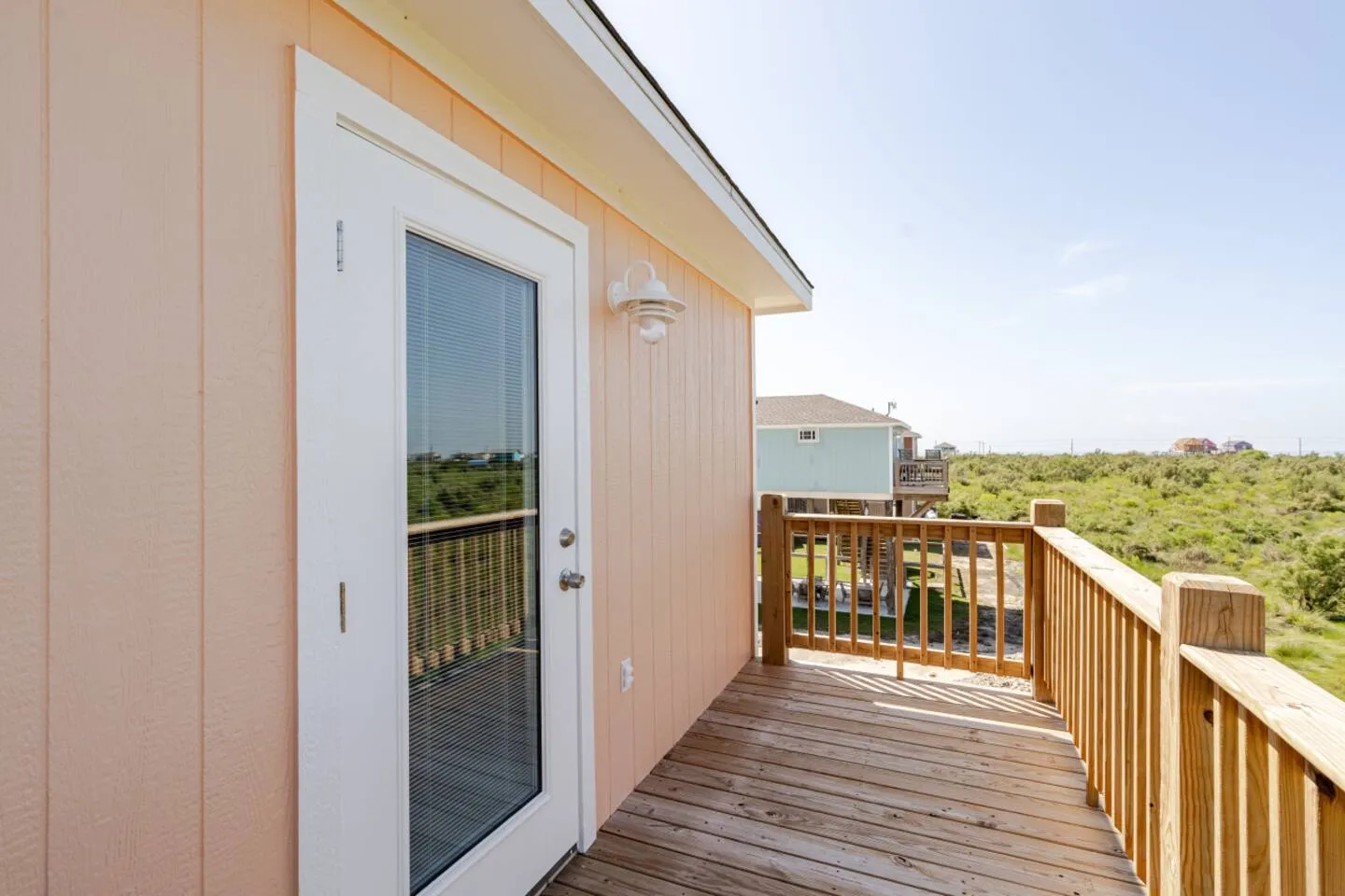 A wooden outdoor deck with wooden railings alongside light peach siding, a white exterior door with glass, and a view of surrounding greenery.