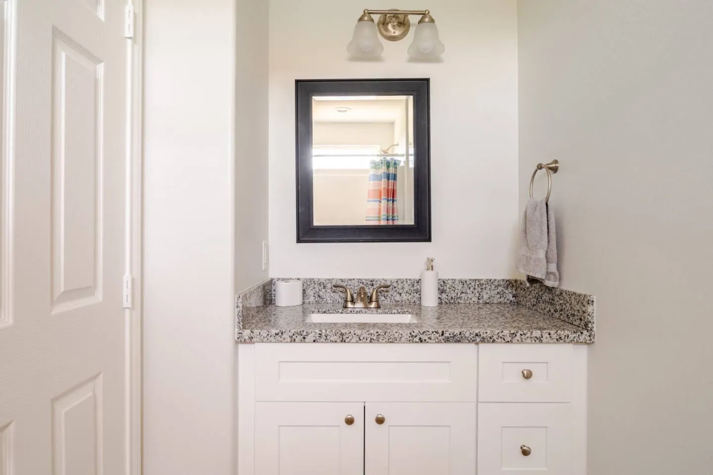 Bathroom vanity with a speckled countertop, undermount sink, white cabinets, dark-framed mirror, and two-bulb light fixture.