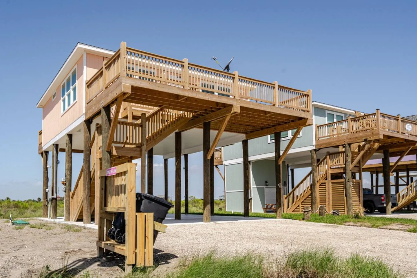 Exterior of an elevated stilt beach home with a large wooden deck and covered ground-level parking underneath.