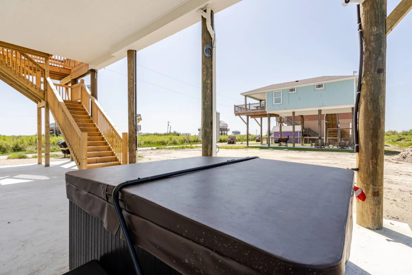 A covered hot tub located on a ground-level concrete slab beneath a stilted house, with wooden stairs leading up and neighboring houses in the background.