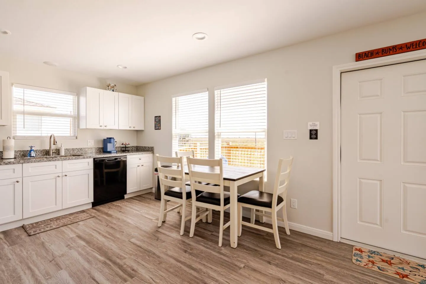 Dining area with a wooden table and white chairs next to kitchen counters and an exterior door.