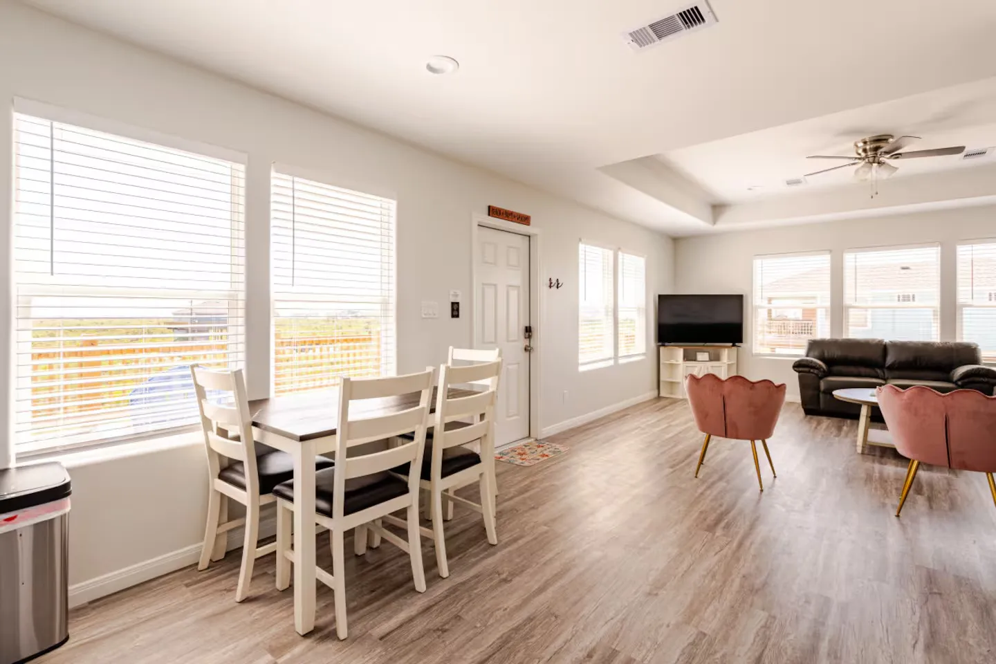Open-concept living and dining area with light wood-style flooring, a dining table with four chairs, pink accent chairs, and a living area in the background.