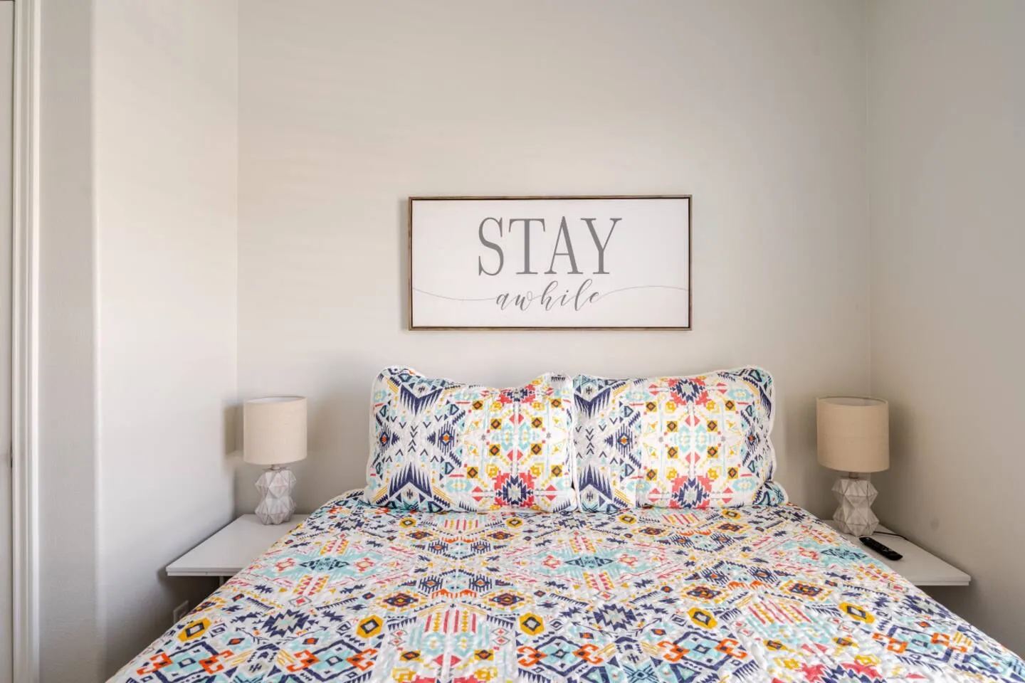 Bedroom featuring a bed with a colorful geometric patterned quilt and matching pillows, flanked by two nightstands with lamps and framed wall art reading Stay awhile.