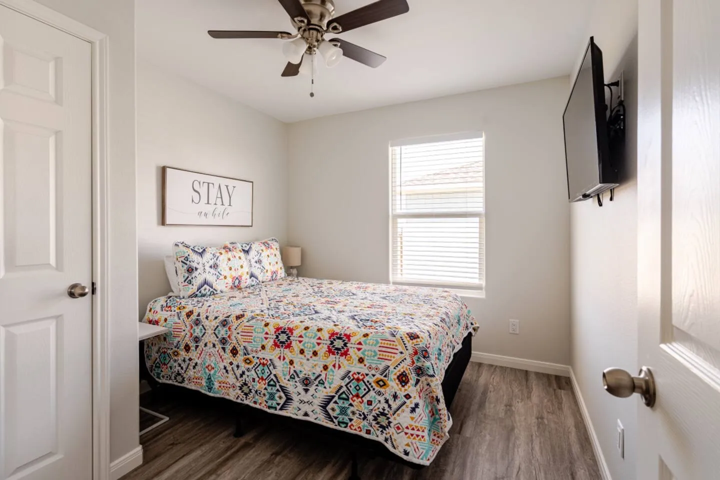 Bedroom with a colorful patterned quilt on the bed, wood-style flooring, a ceiling fan, and a wall-mounted TV.