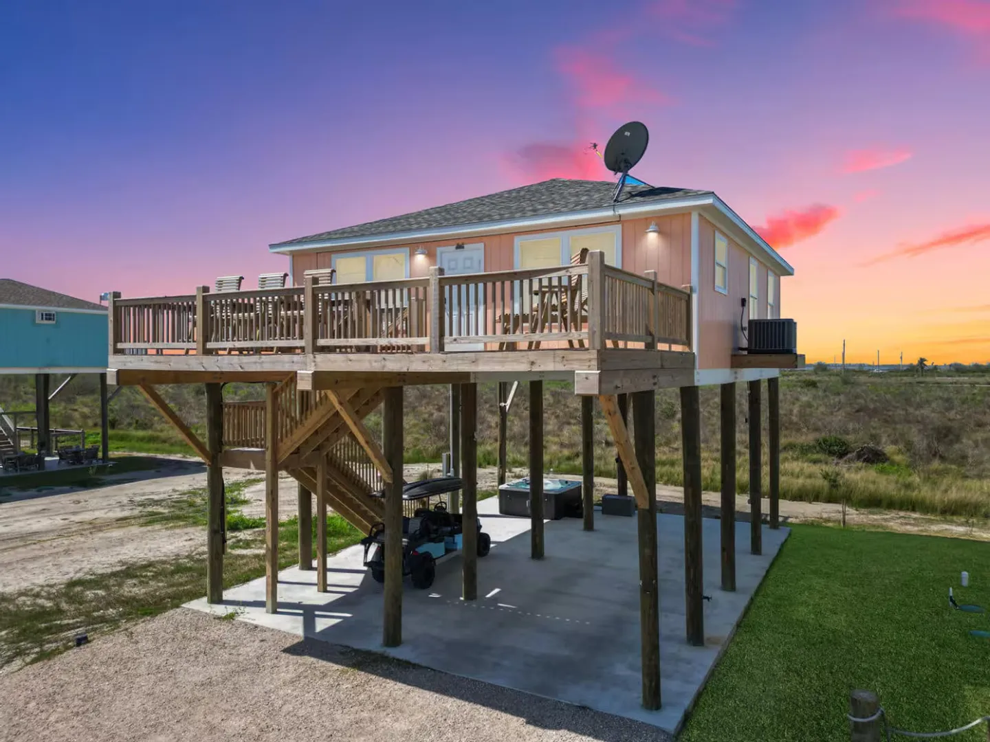 Exterior of an elevated beach house on wooden pilings featuring a large upper deck and a hot tub on the ground-level concrete slab at sunset.