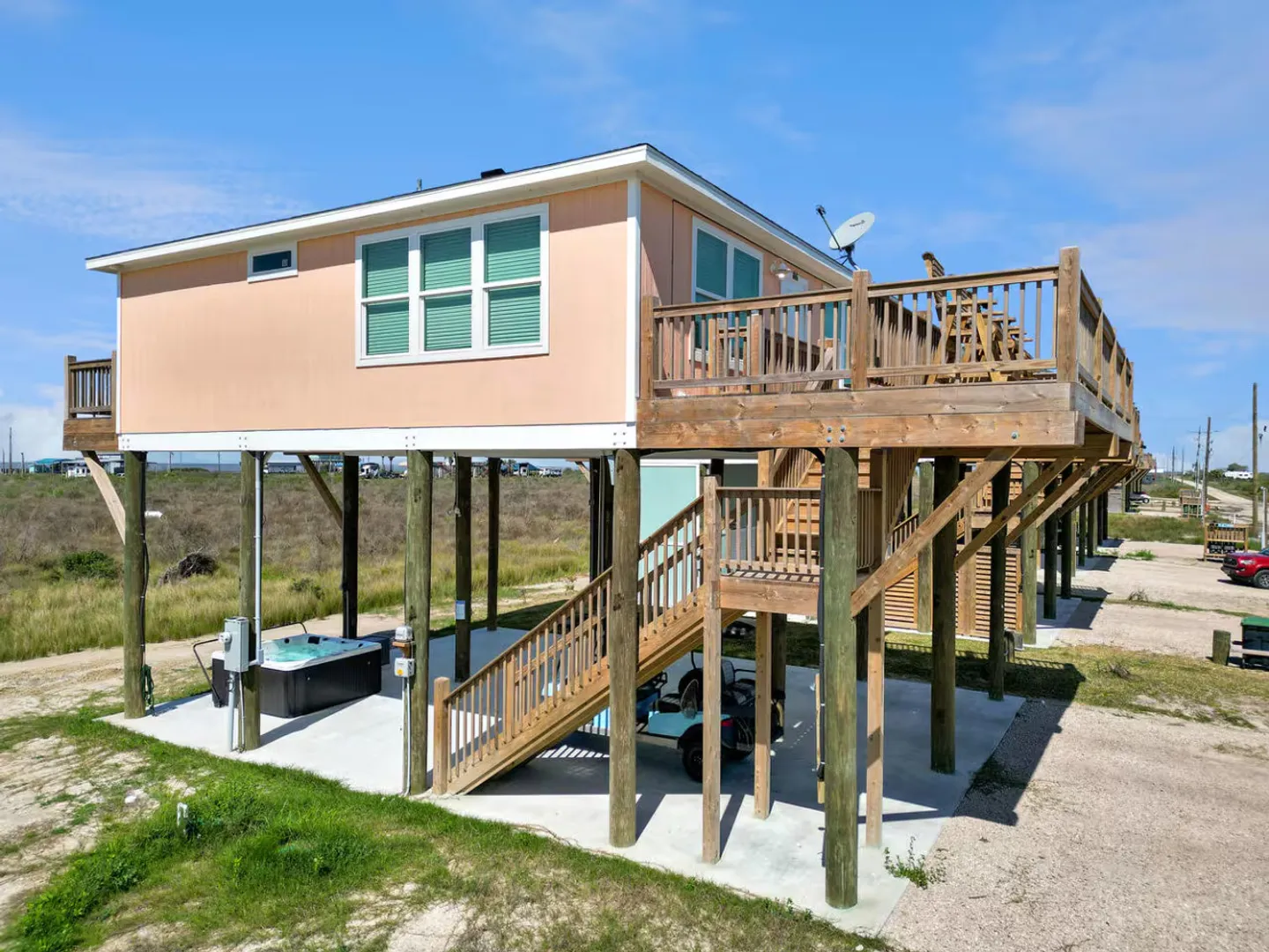 Exterior view of an elevated peach stilt house with an upper deck, wooden stairs, and a ground-level concrete pad featuring a hot tub and golf cart.